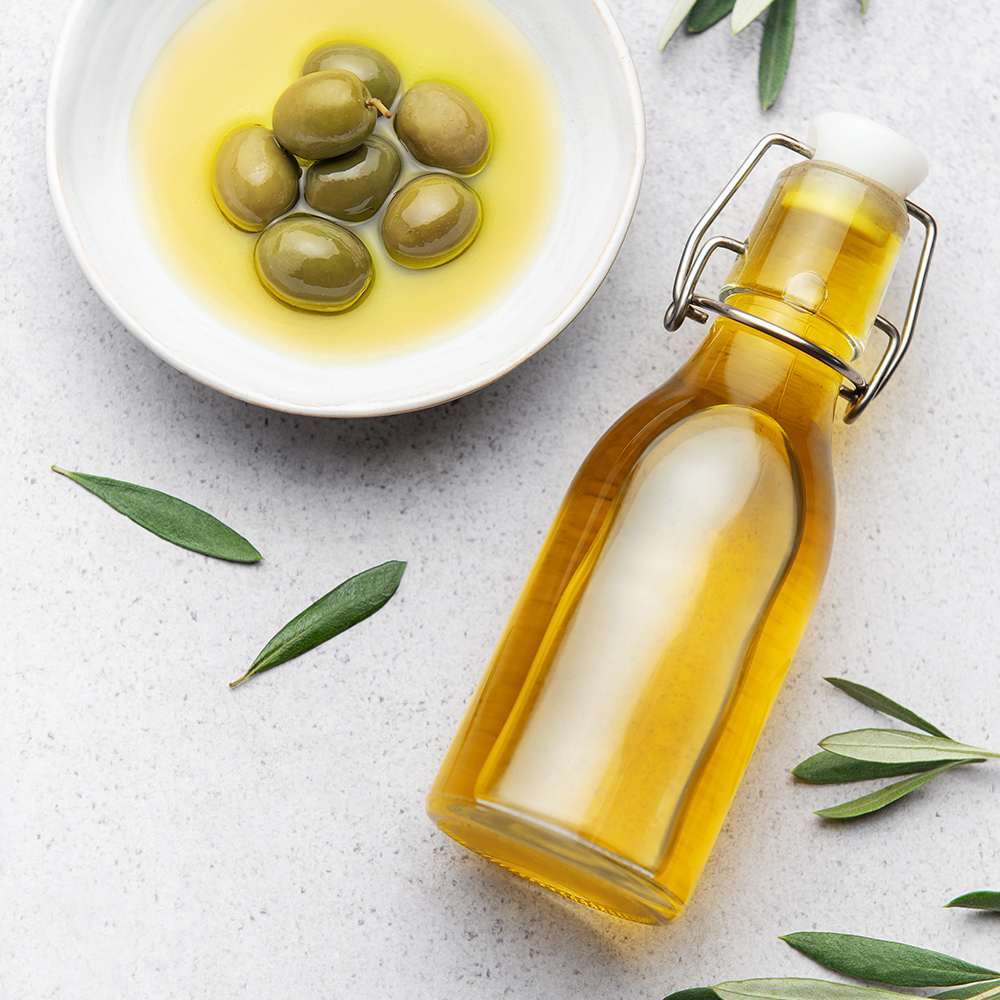 Vista superior de una botella de vidrio con aceite de oliva virgen extra, al lado hay un bowl con aceitunas. Alrededor hay hojas de olivo. Todo sobre un fondo claro.