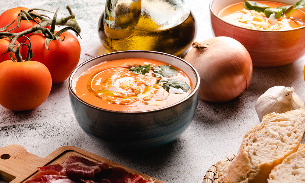 Sopa de tomate en un bowl con especias, acompañada de panes, tomates, aceite y otro bowl con sopa de tomate.