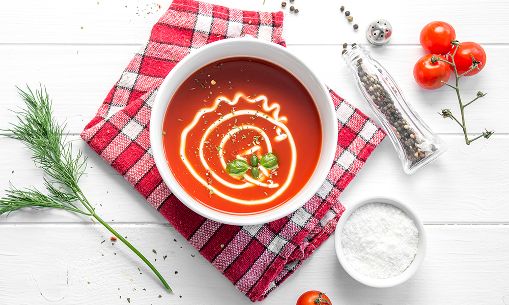 Vista superior de una sopa de tomate en un plato hondo acompañada de albahaca, mayonesa, sal y pimienta sobre una servilleta de cuadros. Todo sobre un fondo blanco.