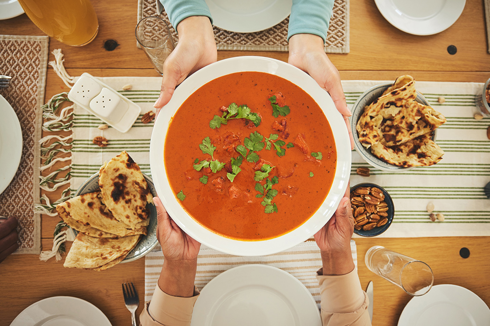 Vista superior de unas manos sosteniendo un plato hondo con una sopa de tomate con especias, de fondo hay una mesa con panes y otros alimentos y platos.