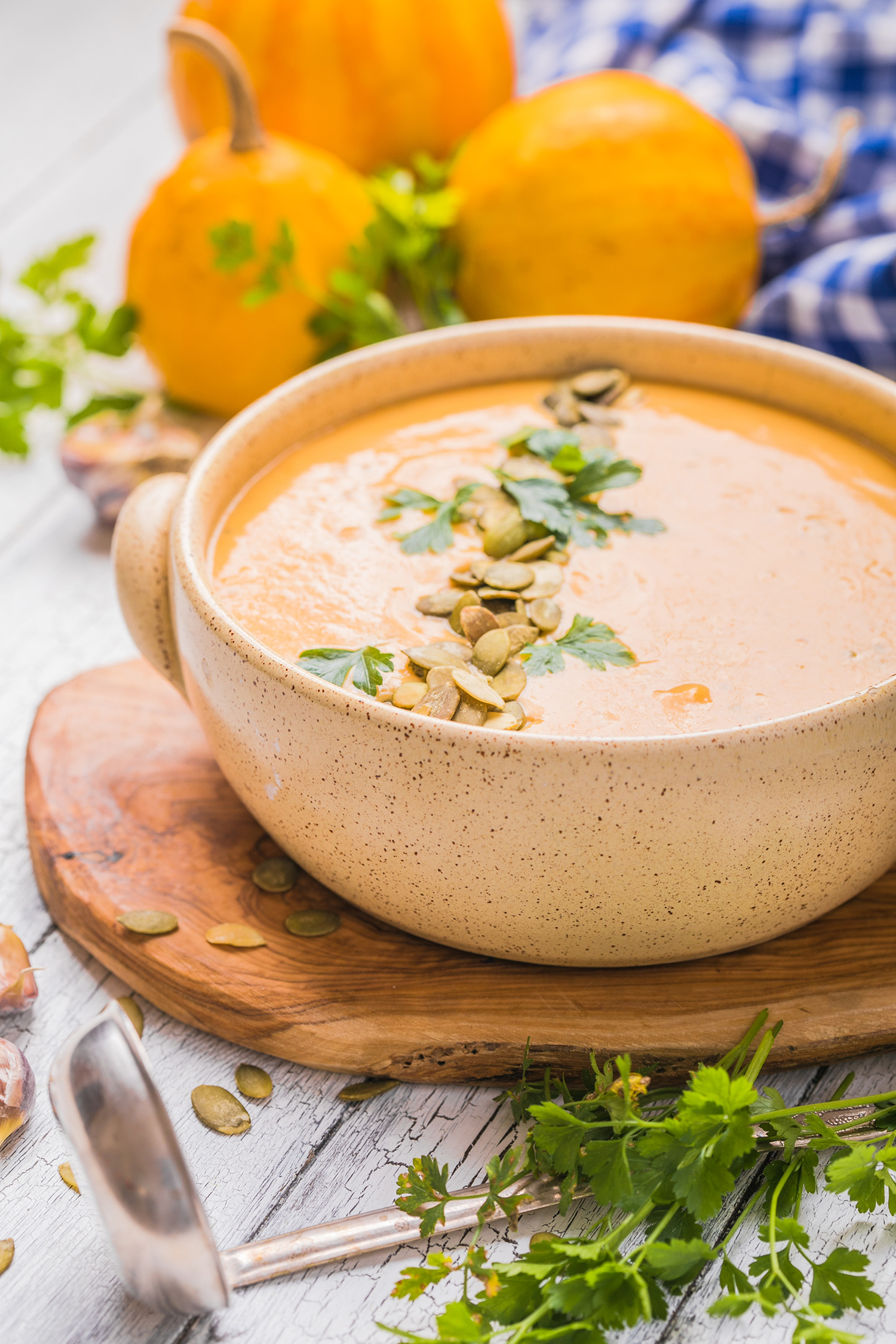 Crema de zanahoria y calabaza en un plato hondo con semillas y especias, acompañado de calabazas pequeñas un cucharón y especias. Todo sobre una mesa clara de madera.