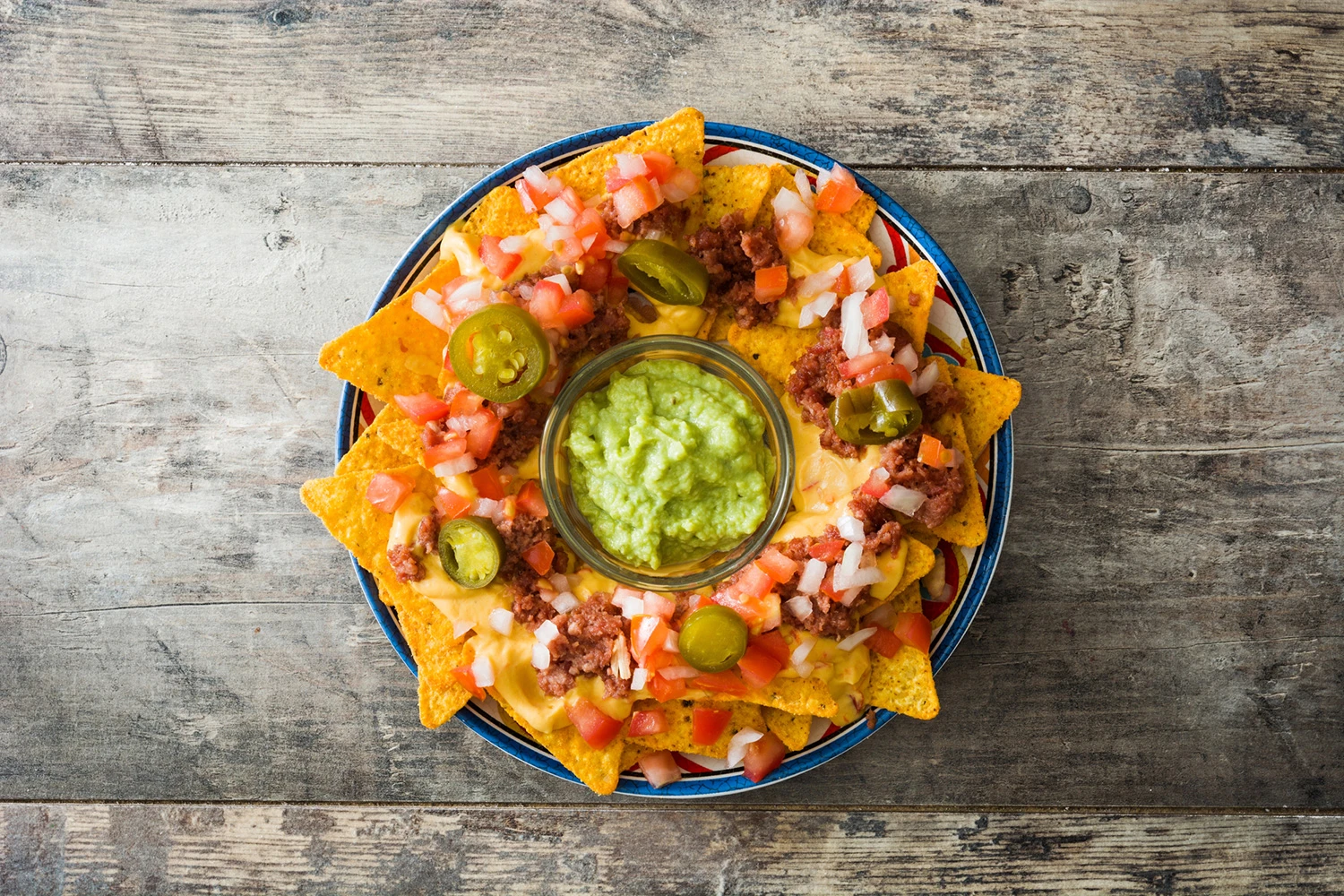 Vista superior de un plato de nachos con cebolla, tomate y jalapeños, acompañado de un mini bowl con guacamole. Todo sobre una mesa oscura de madera.