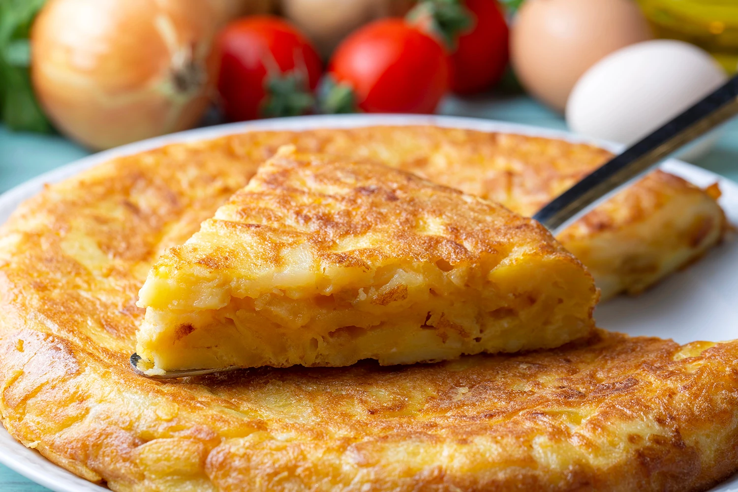 Vista de tortilla española porcionada sobre un plato amplio blanco. Al fondo se aprecian algunos ingredientes crudos, como huevo, cebolla y tomate