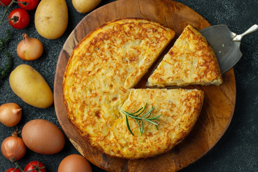 Vista superior de tortilla española sobre un plato de madera. La acompañan ingredientes crudos, como tomate, papa, huevo, ajo y especias, todo sobre una superficie oscura