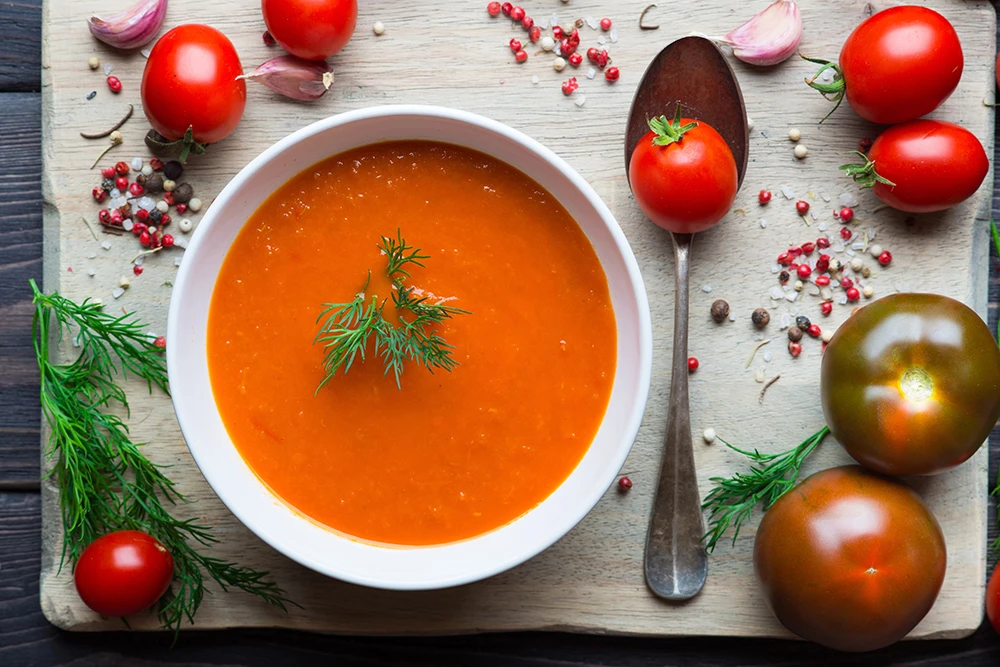 Foto de gazpacho en plato blanco y con ingredientes ubicados a su alrededor, como tomates, pimienta entera y romero. Todo sobre una mesa de madera.