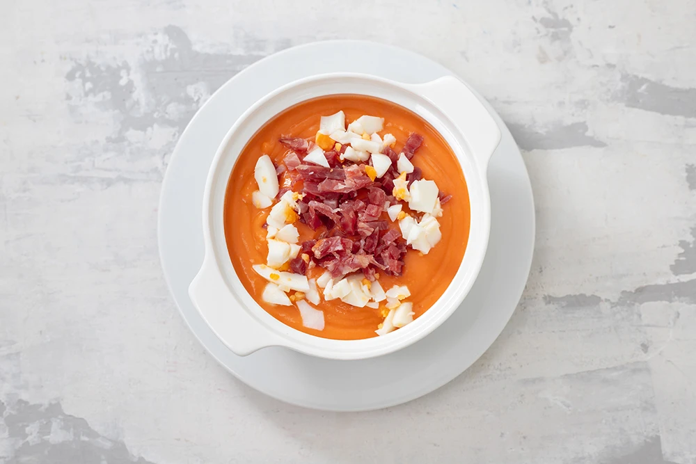 Vista superior de gazpacho servido en plato blanco sobre superficie blanca con gris, encima del gazpacho hay trozos de jamón pequeños.