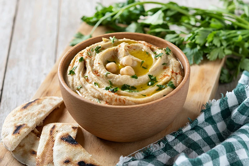 Foto de Hummus de garbanzos en un plato de madera claro, decorado con hojas de perejil, un chorrito de aceite, garbanzos como toppings. Al rededor hay pedazos de tortilla cortados para acompañar y hojas de perejil. Todo sobre una mesa de madera y un mantel a cuadros color verde con blanco.