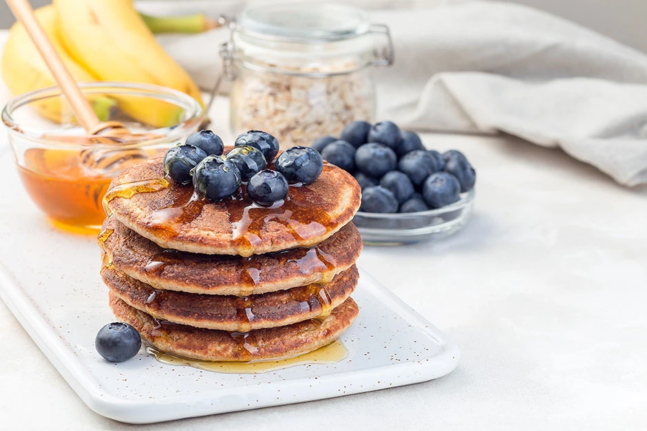 Pancakes de avena apilados en un plato blanco, acompañados de arándanos frescos y miel, sobre una mesa clara con ingredientes de desayuno al fondo.