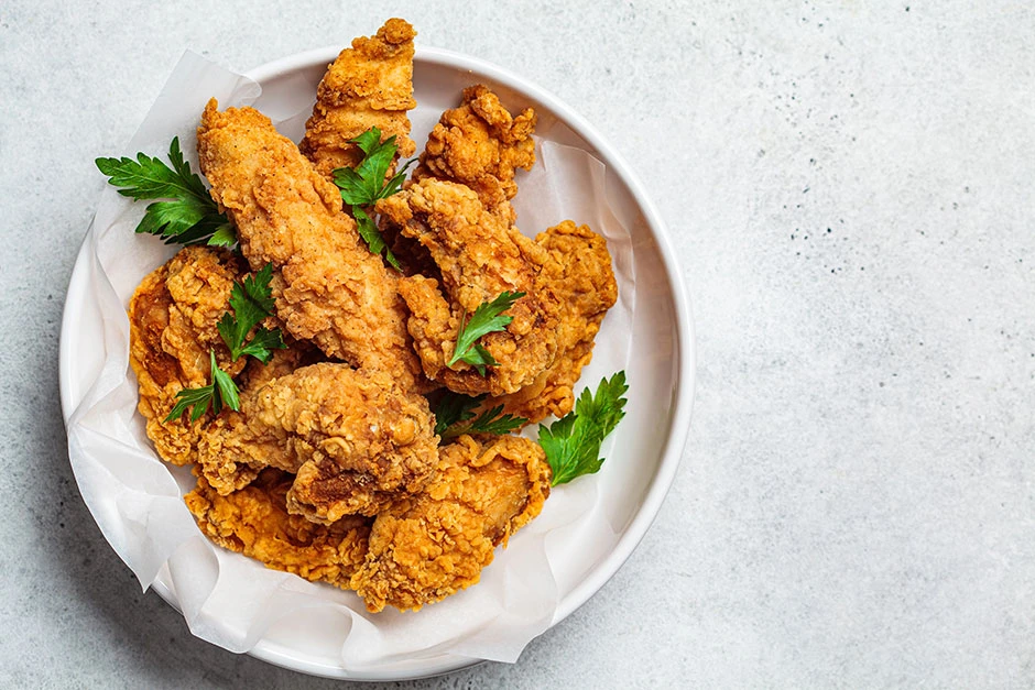 Piezas de pollo frito dorado y crujiente servidas en un plato con hojas de perejil sobre una superficie clara.