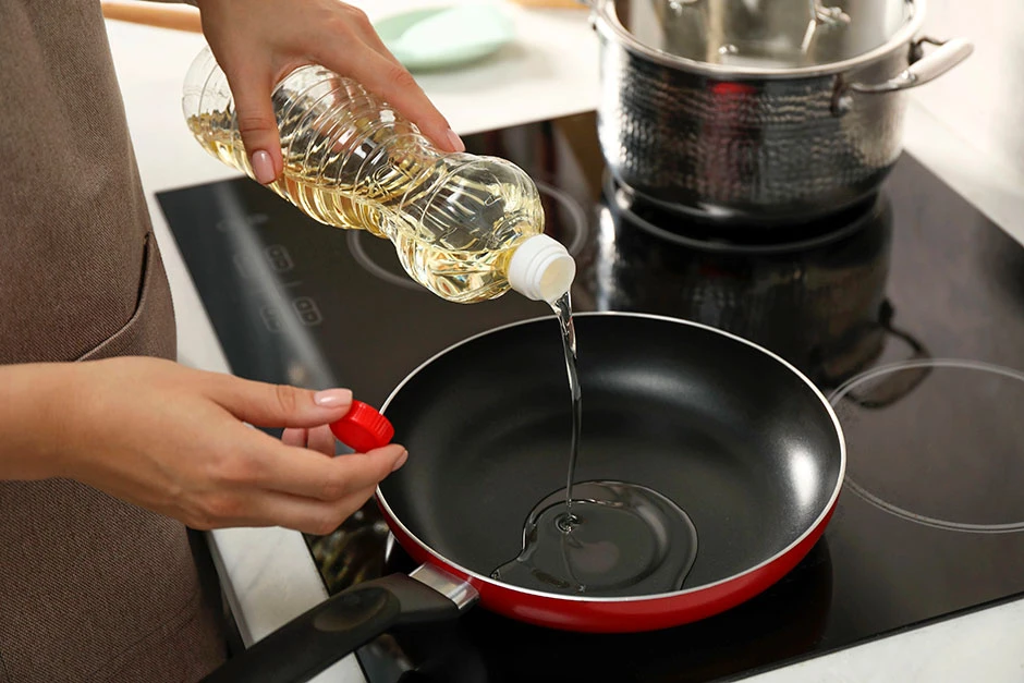 Persona agregando aceite vegetal desde una botella a una sartén sobre una estufa de cocina para preparar una receta.