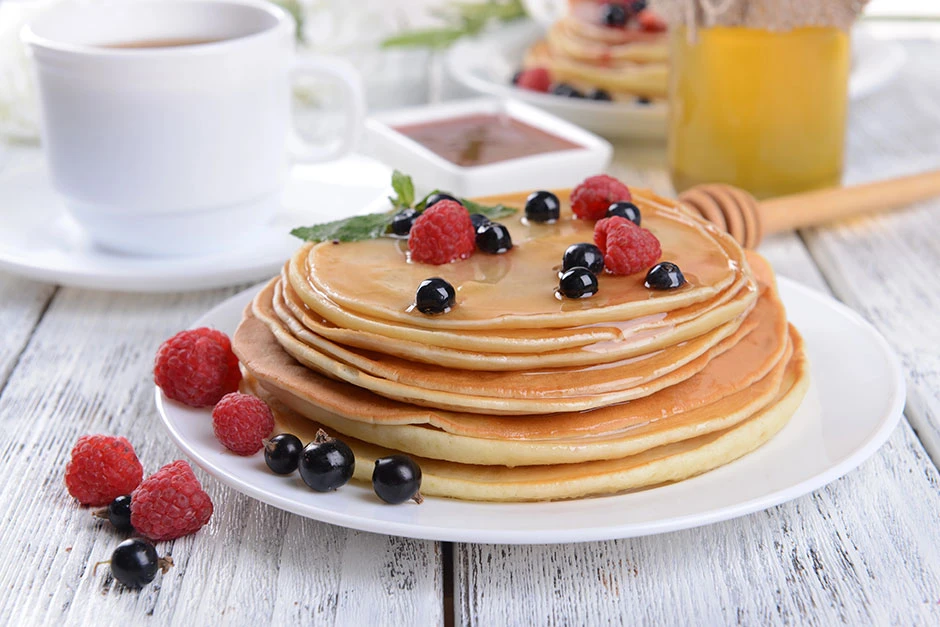 Torre de pancakes caseros con frutas frescas como frambuesas y arándanos, acompañados de miel y una taza de café sobre una mesa de madera clara.