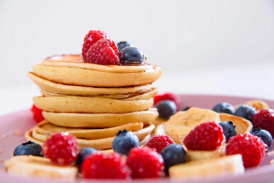Pancakes caseros apilados con frutos rojos frescos alrededor, servidos en un plato sobre una superficie clara con espacio libre para composición.