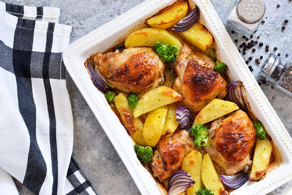 Pollo al horno dorado acompañado de papas en cascos, brócoli y cebolla morada, servido en una refractaria blanca.
