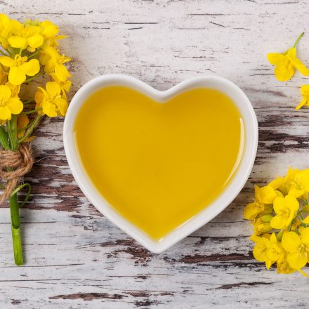Rapeseed oil in heart shaped bowl and flowers on wooden background, top view.